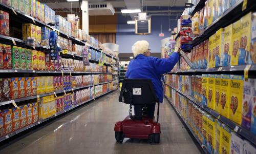 An older woman in a scooter goes down the cereal aisle of a supermarket